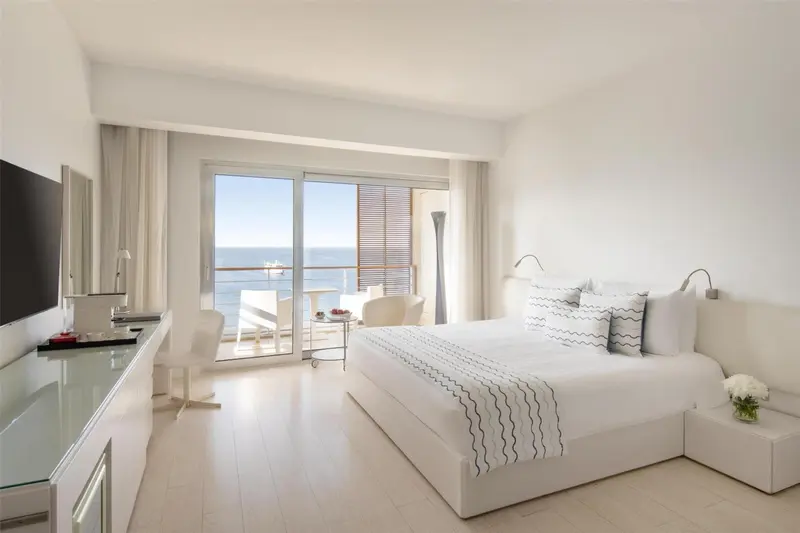 Chambre lumineuse avec un grand lit et une vue sur la mer depuis un balcon.