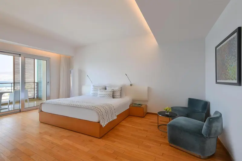Chambre lumineuse avec un grand lit et un coin salon près d'une baie vitrée.