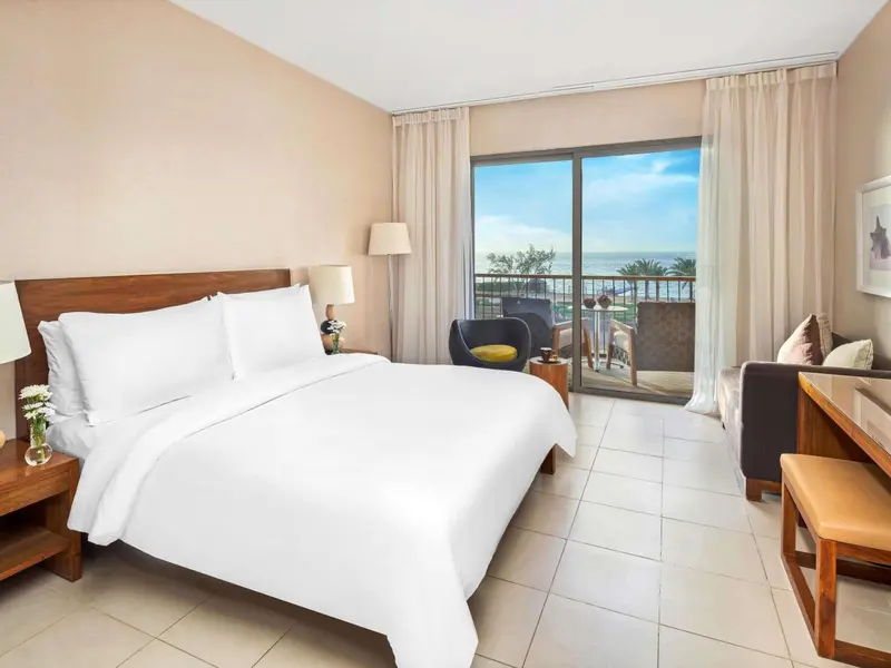Chambre lumineuse avec un grand lit et une vue sur la mer depuis un balcon.