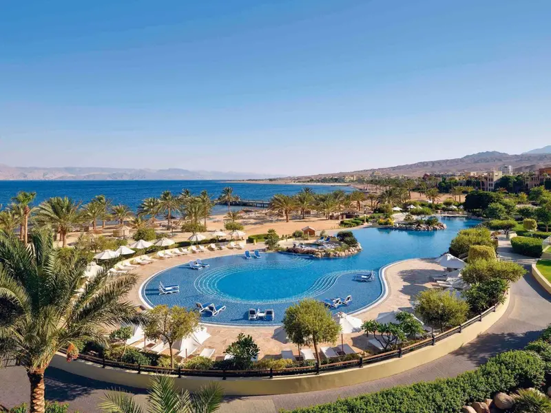 Vue aérienne d'une piscine sinueuse bordée de transats et de palmiers, avec la mer en arrière-plan.