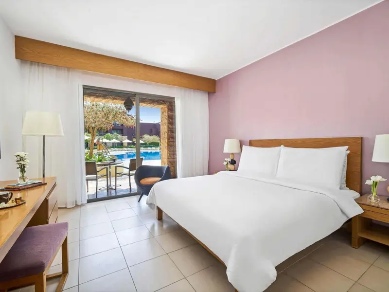 Chambre lumineuse avec un grand lit et vue sur une piscine.