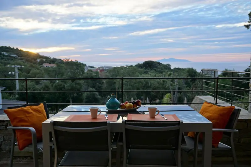 Une table est dressée sur une terrasse avec vue sur un paysage vallonné et la mer.