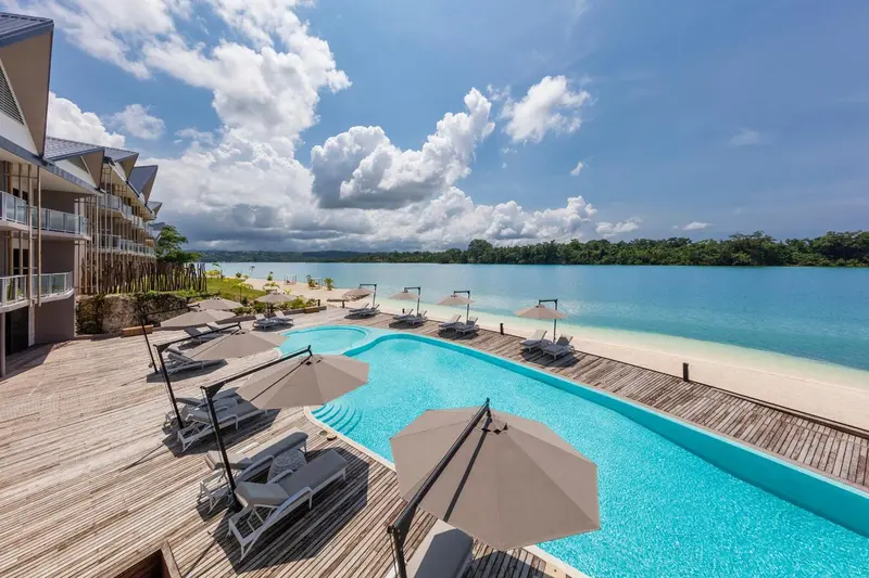 Une piscine turquoise bordée de transats et de parasols, avec une plage de sable fin et une mer bleue en arrière-plan.