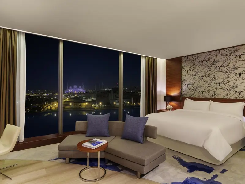 Chambre moderne avec un grand lit et un coin salon face à une vue nocturne sur la ville.