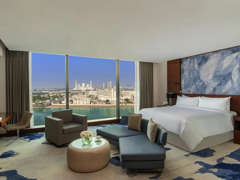 Chambre lumineuse avec un grand lit et un coin salon face à une vue sur la mer et la ville.