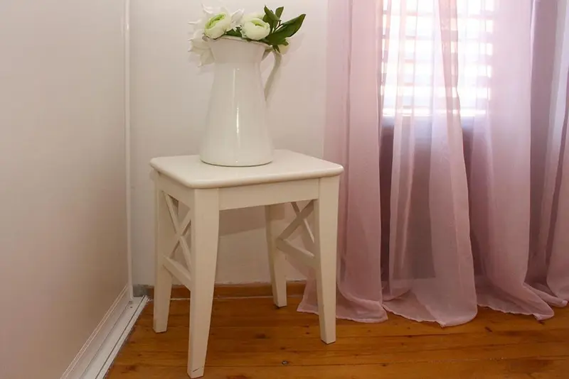 Un vase de fleurs blanches repose sur une petite table blanche près d'une fenêtre.