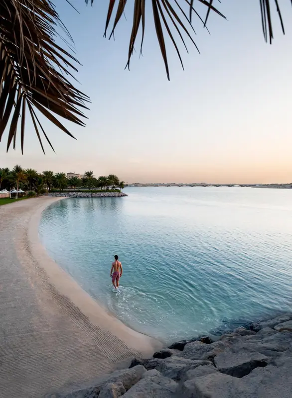 Une personne se tient dans l'eau turquoise le long d'une plage de sable clair au lever du soleil.