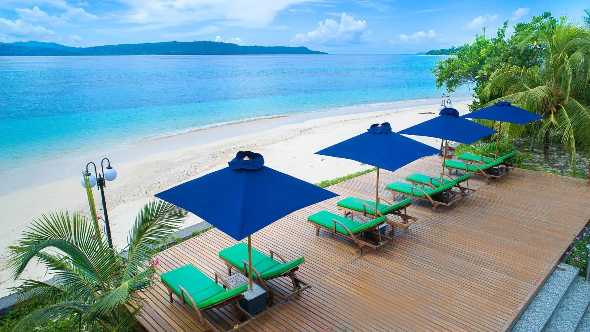 Transats bleus et verts sous des parasols sur une terrasse en bois face à une plage de sable blanc.