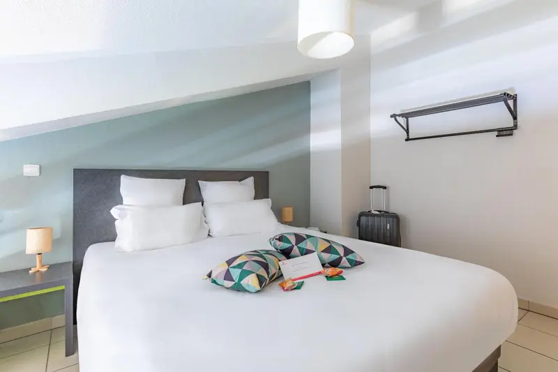 Une chambre lumineuse avec un lit fait et une valise posée au pied.