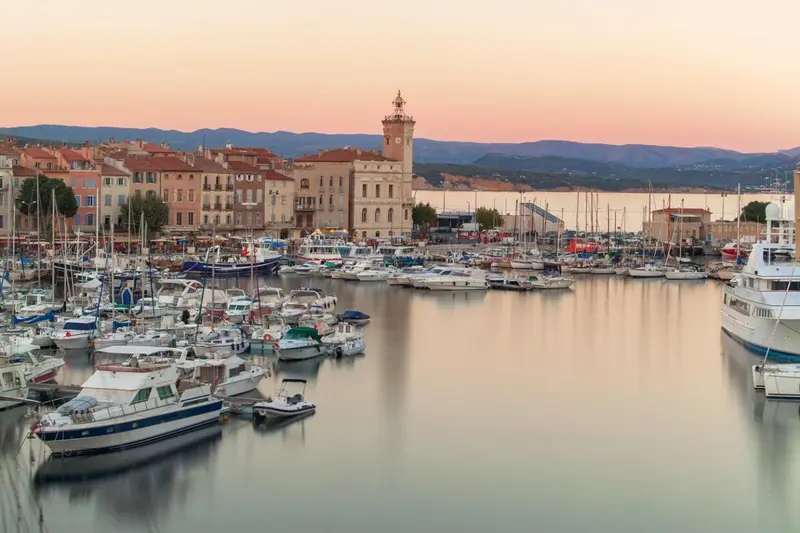 Vue d'un port de plaisance au coucher du soleil avec des bateaux amarrés et une tour en arrière-plan.