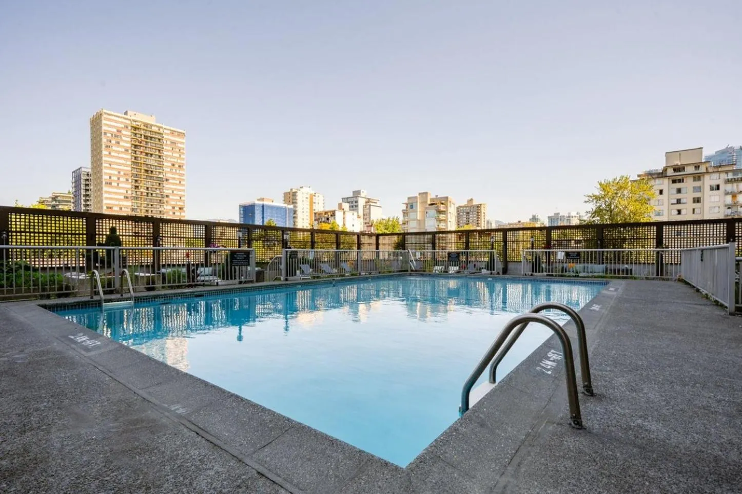 Piscine rectangulaire entourée d'une terrasse en béton avec des transats et des bâtiments en arrière-plan.