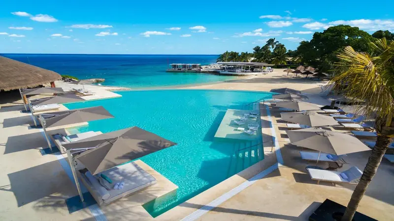 Vue aérienne d'une piscine turquoise bordée de transats sur une plage de sable blanc.