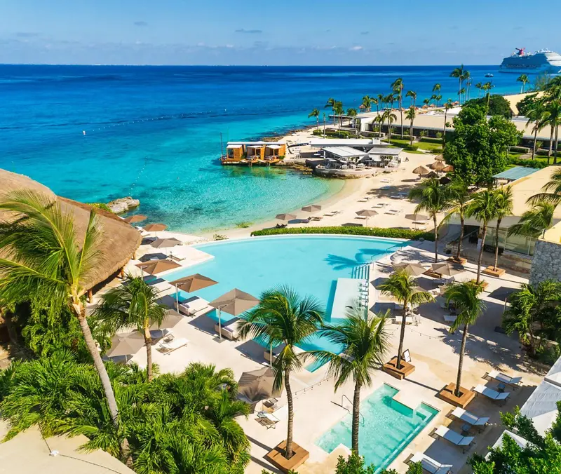Vue aérienne d'une plage avec des transats et des palmiers, bordée d'une piscine et d'une mer turquoise.