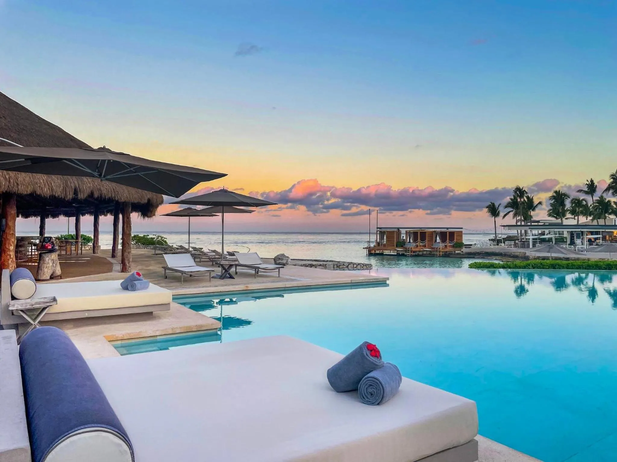 Transats blancs au bord d'une piscine turquoise avec vue sur la mer au coucher du soleil.