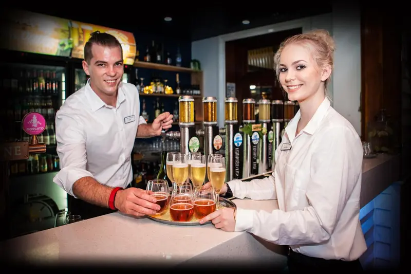 Deux employés souriants servent des boissons derrière un comptoir de bar bien garni.