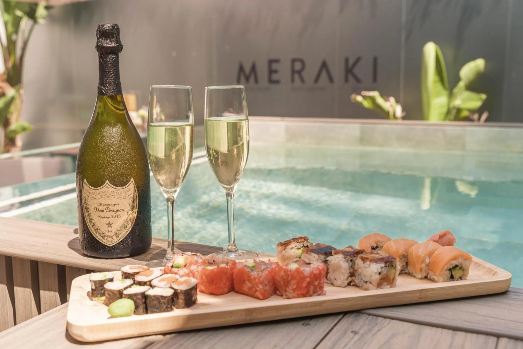 Une bouteille de champagne, deux verres et un assortiment de sushis sont disposés sur une table au bord d'une piscine.