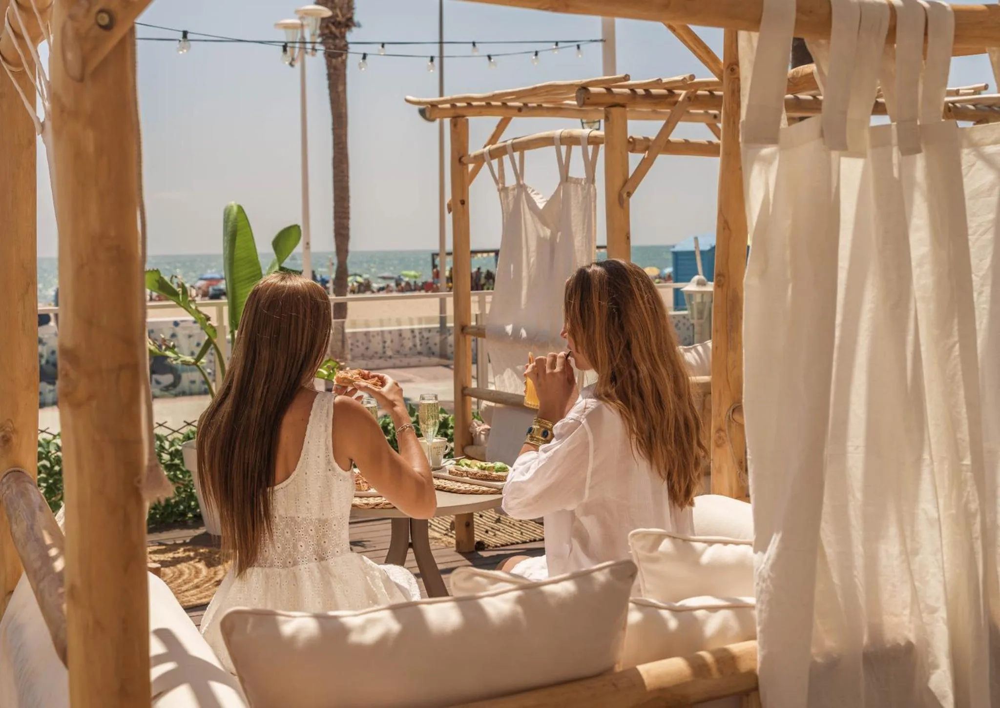Deux femmes déjeunent à une table en bois sous une pergola ombragée en bord de mer.