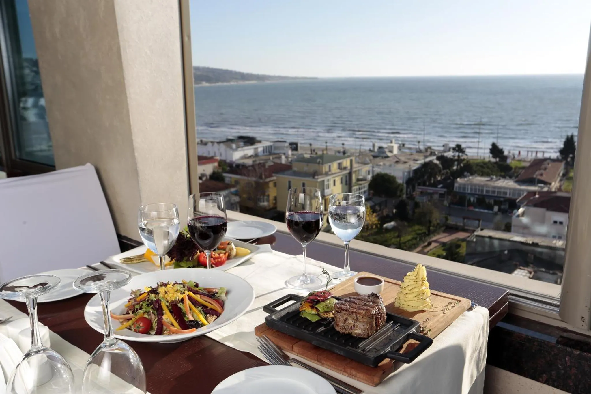 Une table est dressée avec des plats et des verres de vin, offrant une vue sur la mer et la ville.