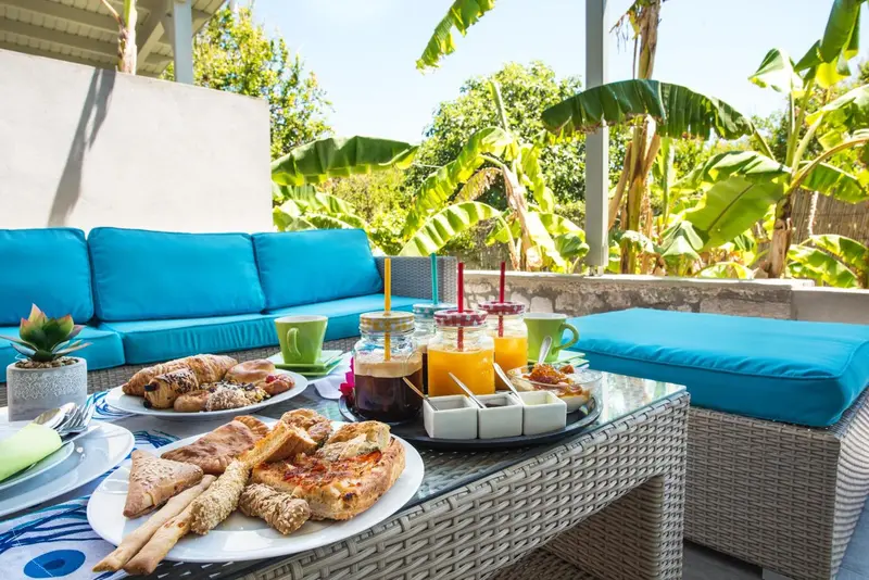 Une table basse en osier présente un petit-déjeuner avec des viennoiseries et des boissons fraîches sur une terrasse.