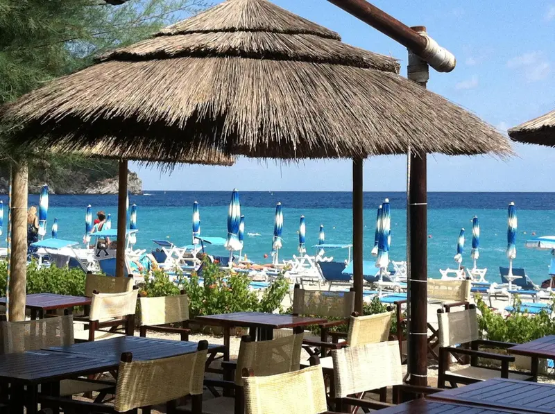 Tables et chaises en osier sous des parasols de chaume face à une plage turquoise.