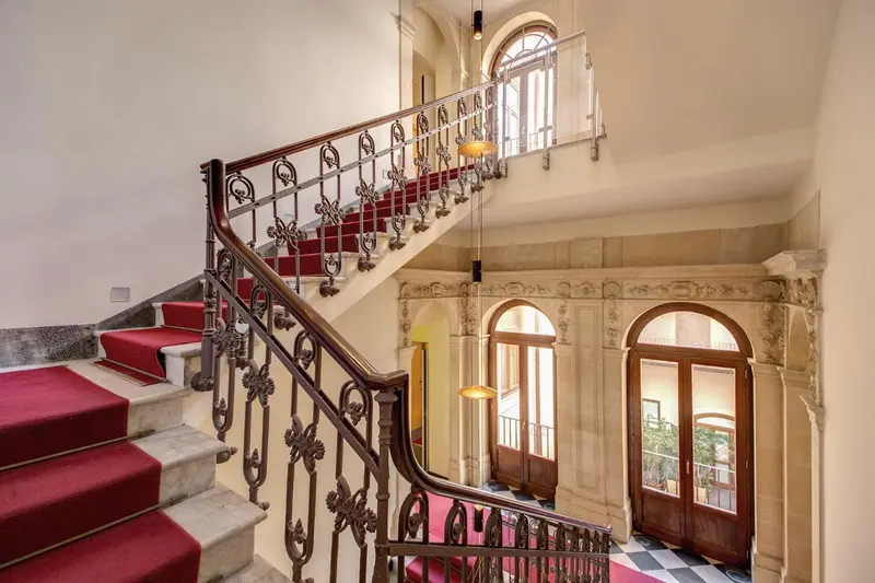 Escalier en pierre avec rampe en fer forgé et tapis rouge dans un hall d'entrée.