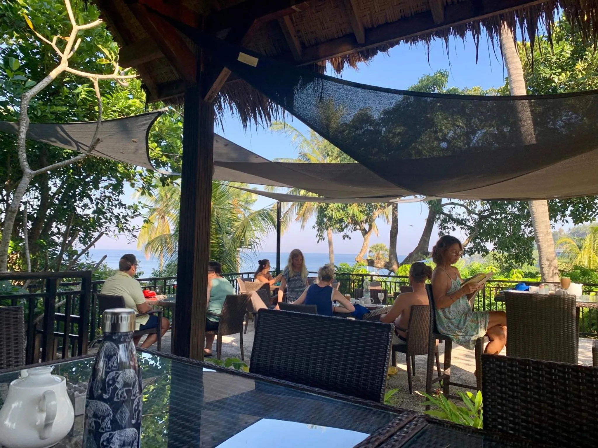 Des clients sont assis à des tables en osier sur une terrasse ombragée avec vue sur la mer.
