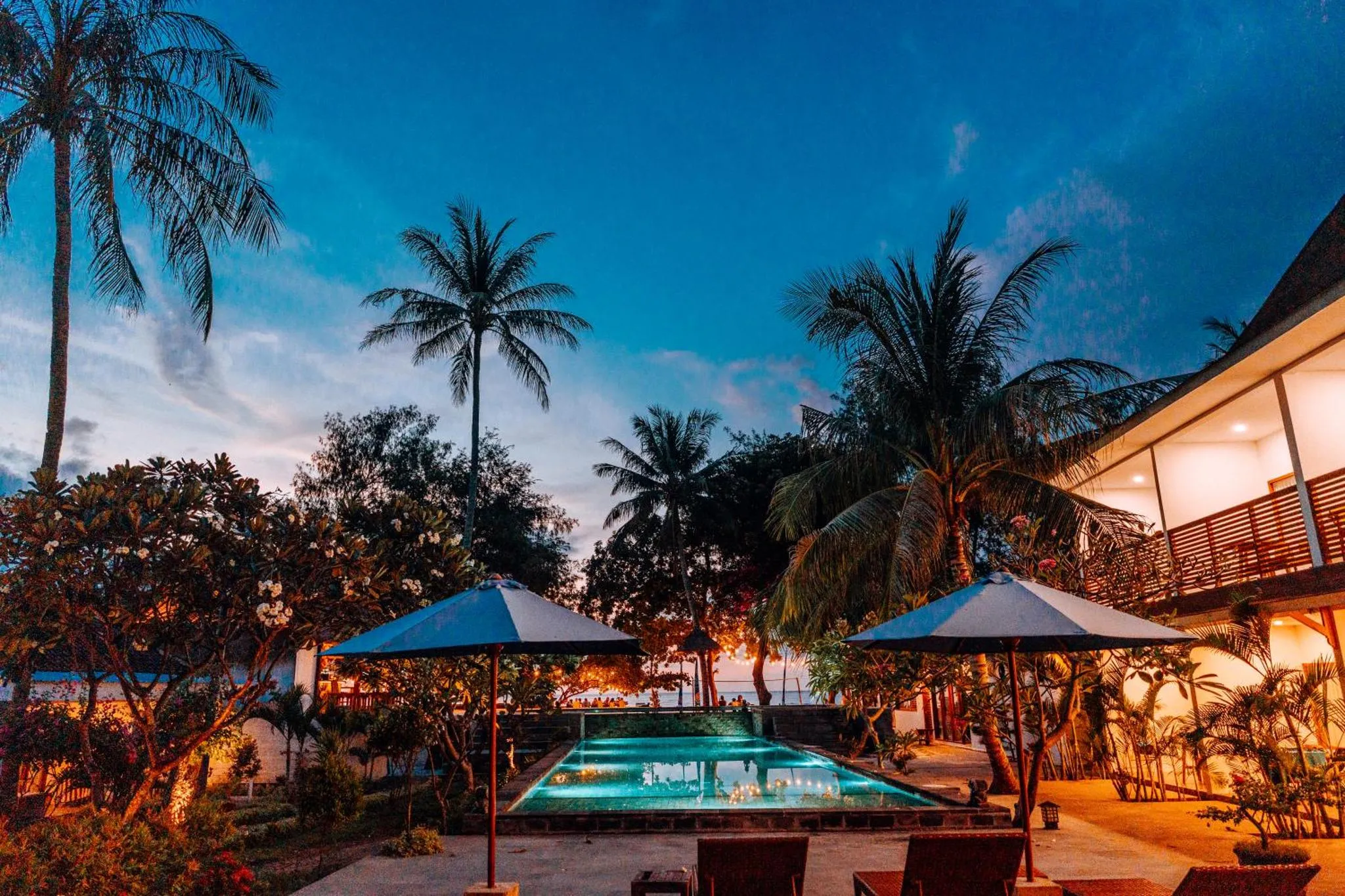 Une piscine éclairée au crépuscule, entourée de végétation tropicale et de quelques parasols.