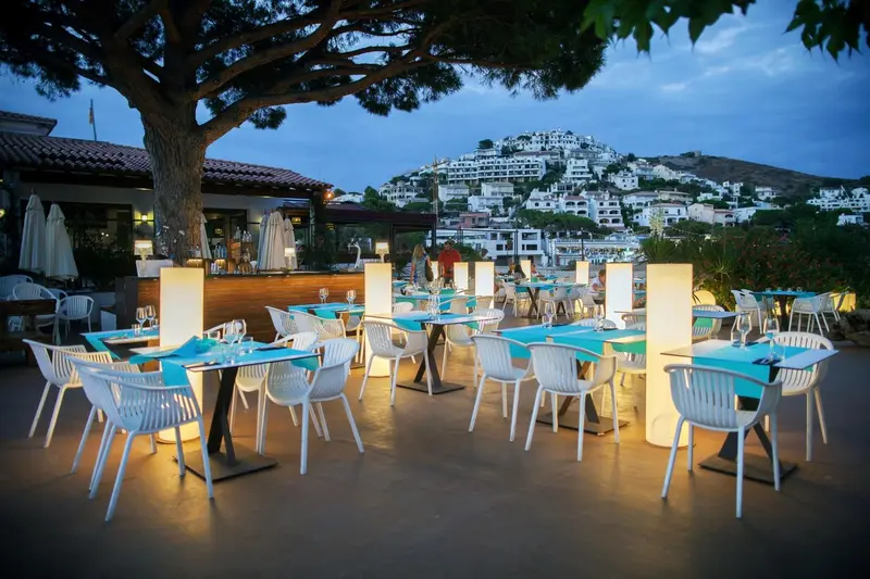 Terrasse lumineuse avec tables et chaises face à une ville côtière.