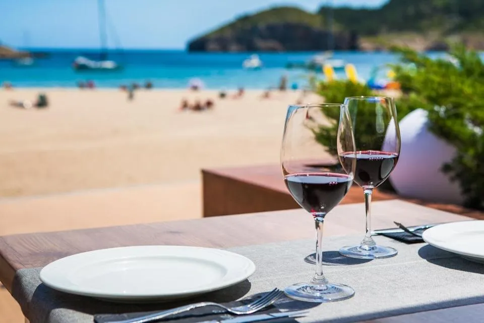 Deux verres de vin rouge et une assiette sur une table en terrasse face à la mer.