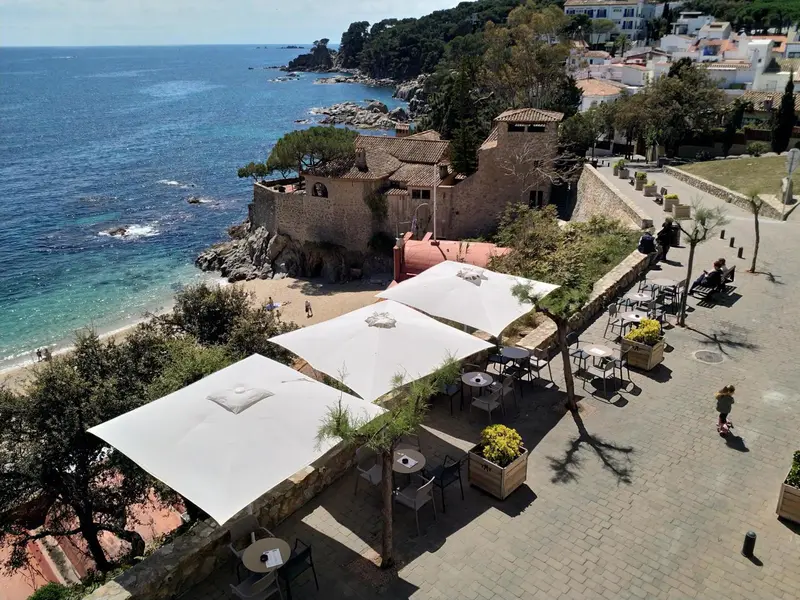 Terrasse ombragée avec tables et chaises surplombant une crique rocheuse.