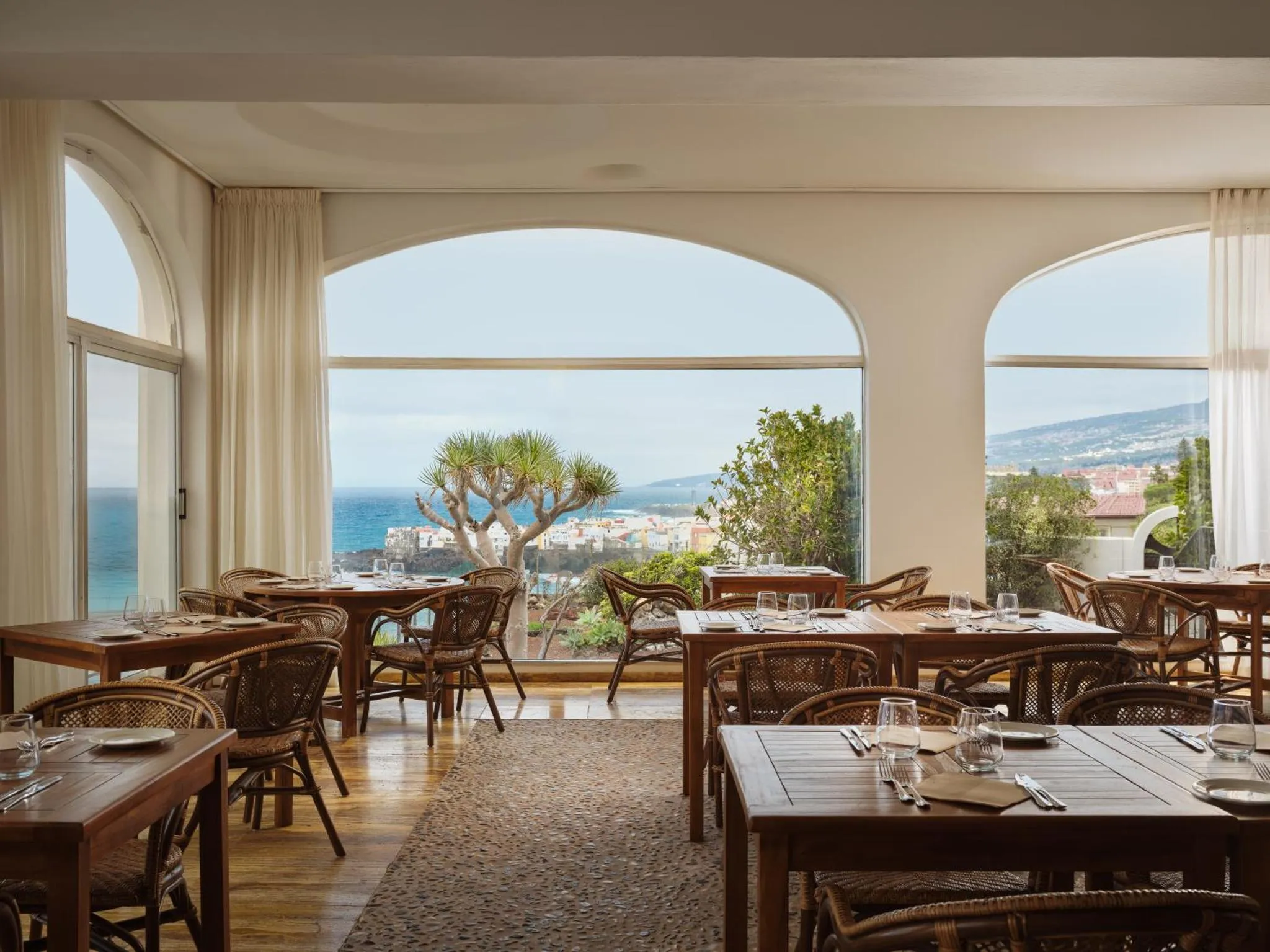 Une salle lumineuse avec des tables dressées face à une vue sur la mer et les collines.