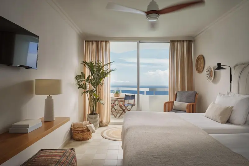 Chambre lumineuse avec un lit fait et vue sur la mer depuis un balcon.