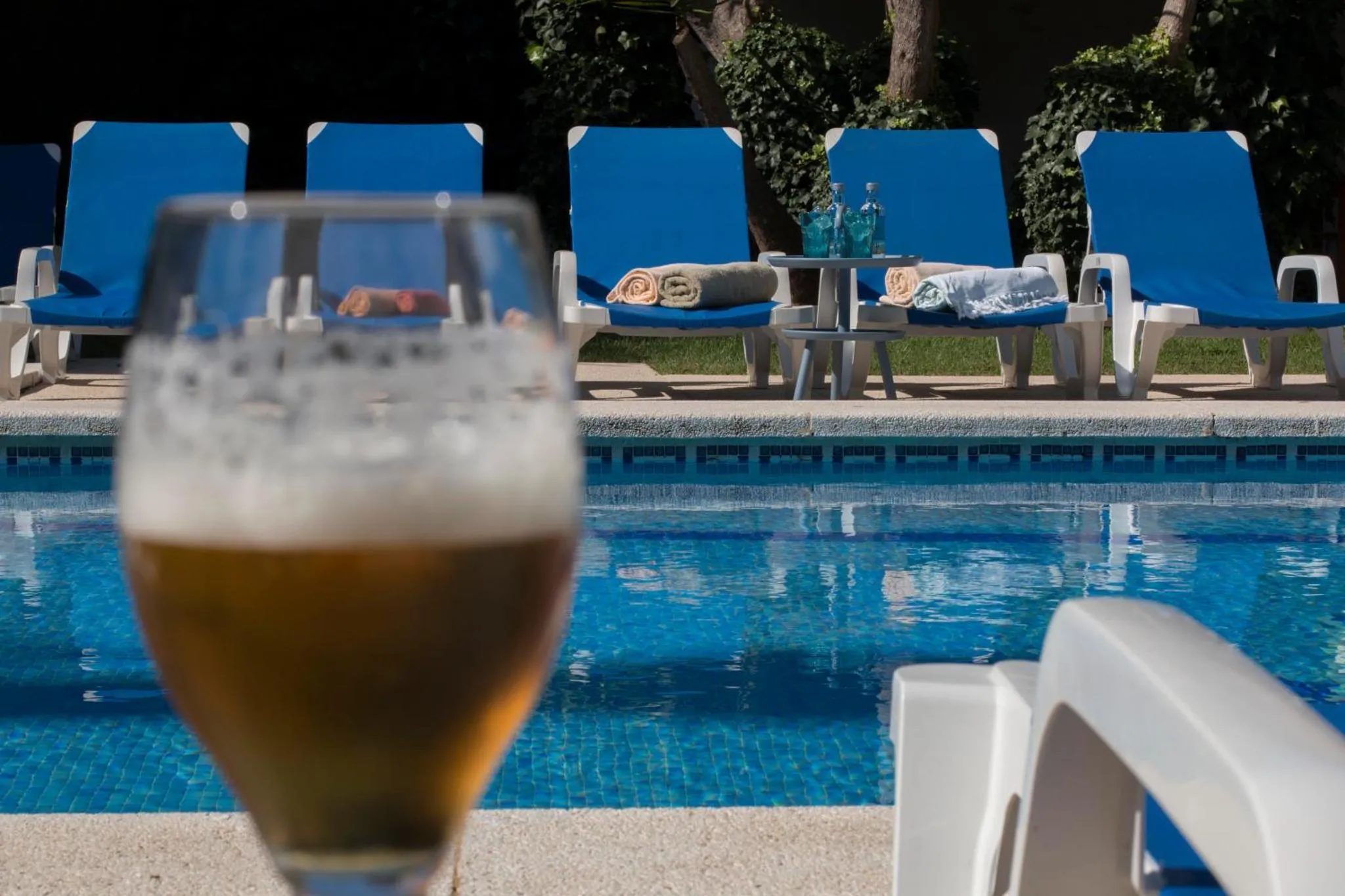 Un verre de bière au premier plan, avec des transats bleus et une piscine en arrière-plan.