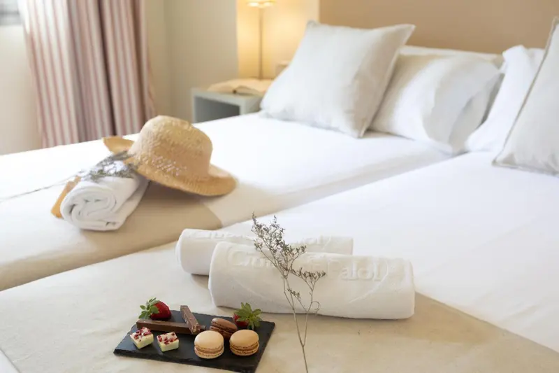 Un lit recouvert de draps blancs et d'oreillers, avec des serviettes et des gourmandises sur la table de chevet.