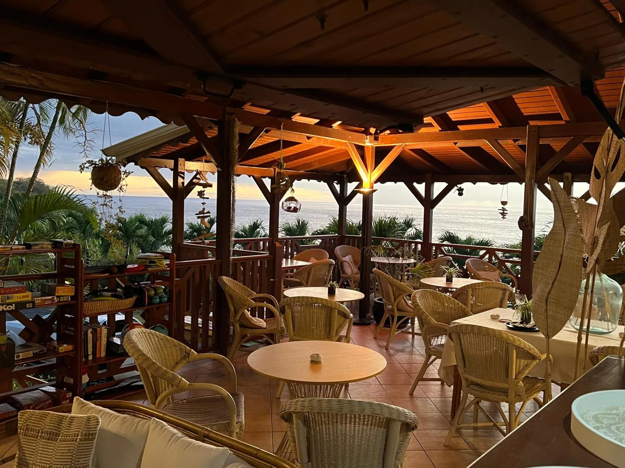 Terrasse ombragée avec tables et chaises face à la mer au coucher du soleil.