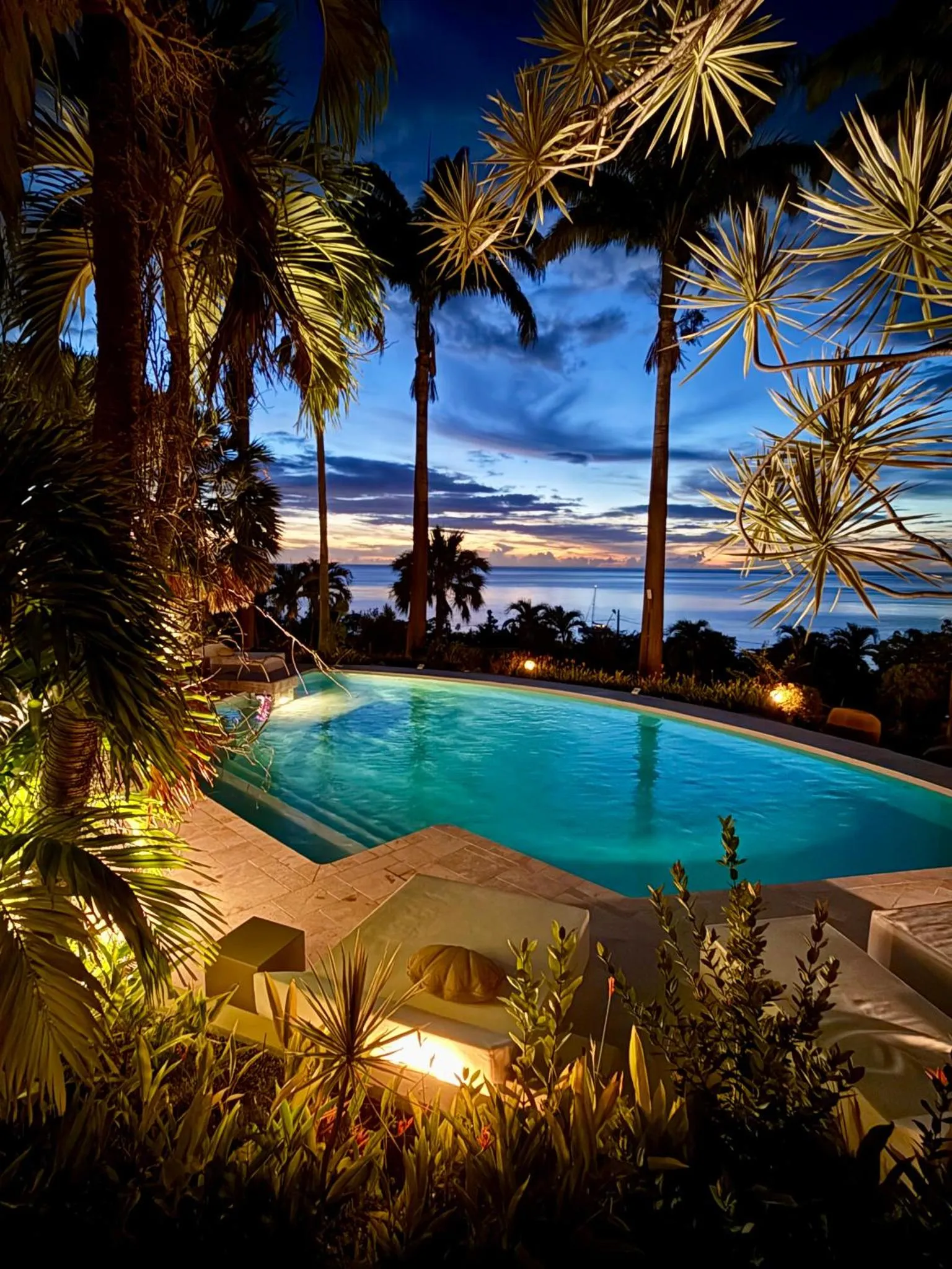 Une piscine illuminée au crépuscule, entourée de végétation tropicale et surplombant la mer.