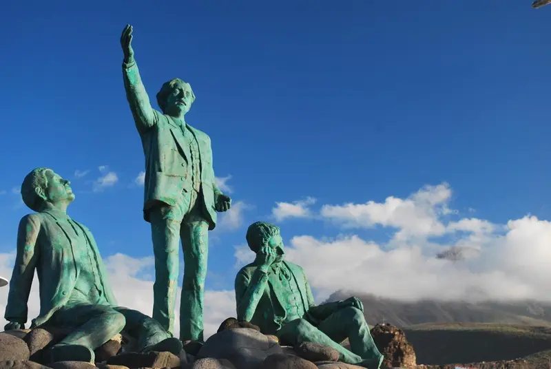 Quatre statues de personnages en bronze regardent vers l'horizon sous un ciel bleu nuageux.