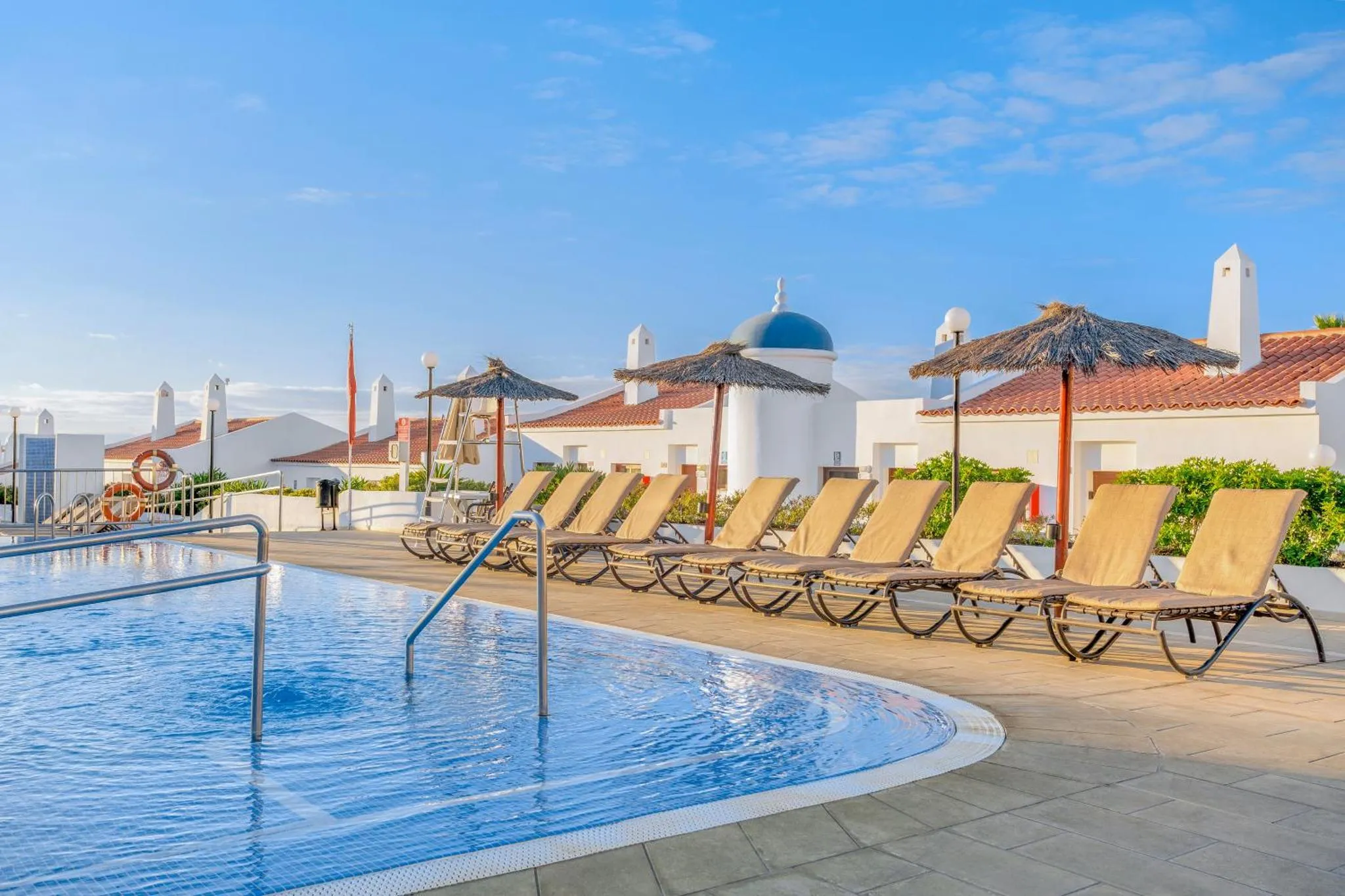 Transats alignés au bord d'une piscine ensoleillée avec des parasols en chaume.