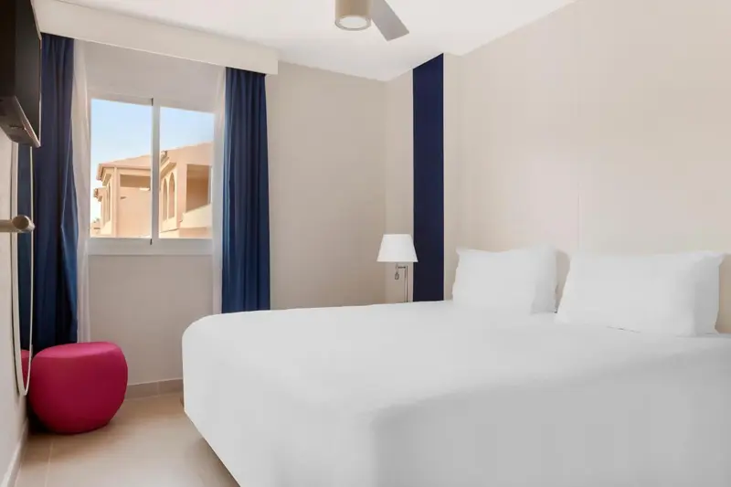 Une chambre lumineuse avec un lit blanc et un pouf rose.
