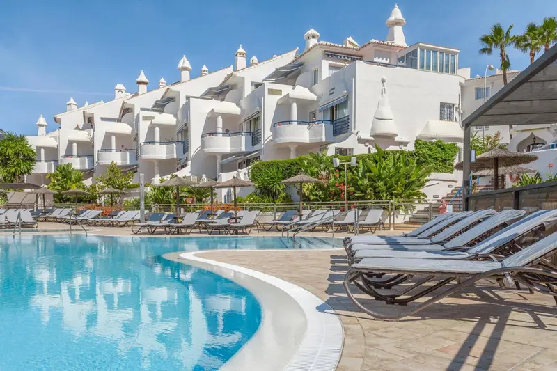 Transats alignés au bord d'une piscine turquoise devant un bâtiment blanc.