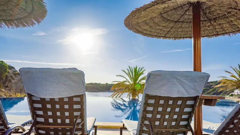 Deux transats blancs sont placés au bord d'une piscine turquoise sous des parasols en chaume.