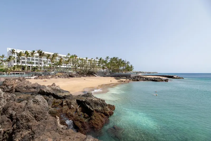 Vue d'une plage de sable bordée de rochers volcaniques et d'un bâtiment blanc.