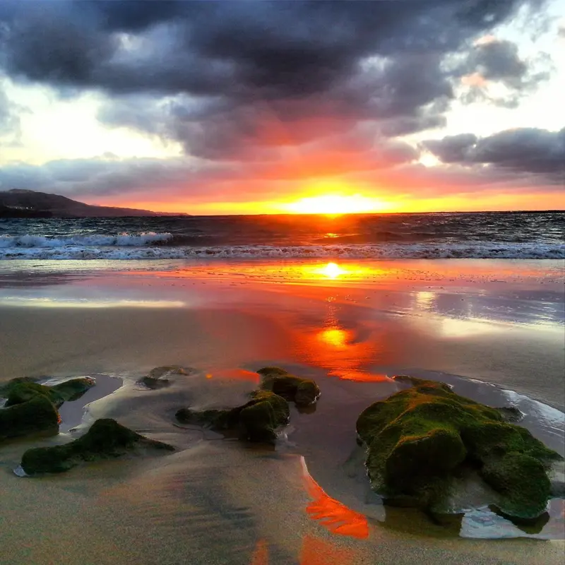Coucher de soleil sur une plage de sable avec des rochers au premier plan.