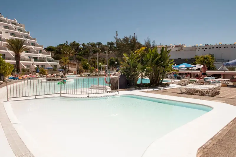 Piscine ensoleillée avec transats et végétation luxuriante devant un bâtiment blanc.