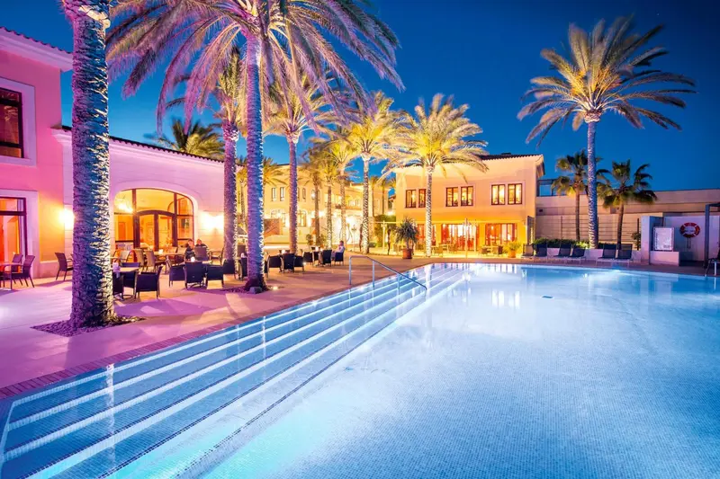 Une piscine illuminée au premier plan, avec un bâtiment et des palmiers en arrière-plan.