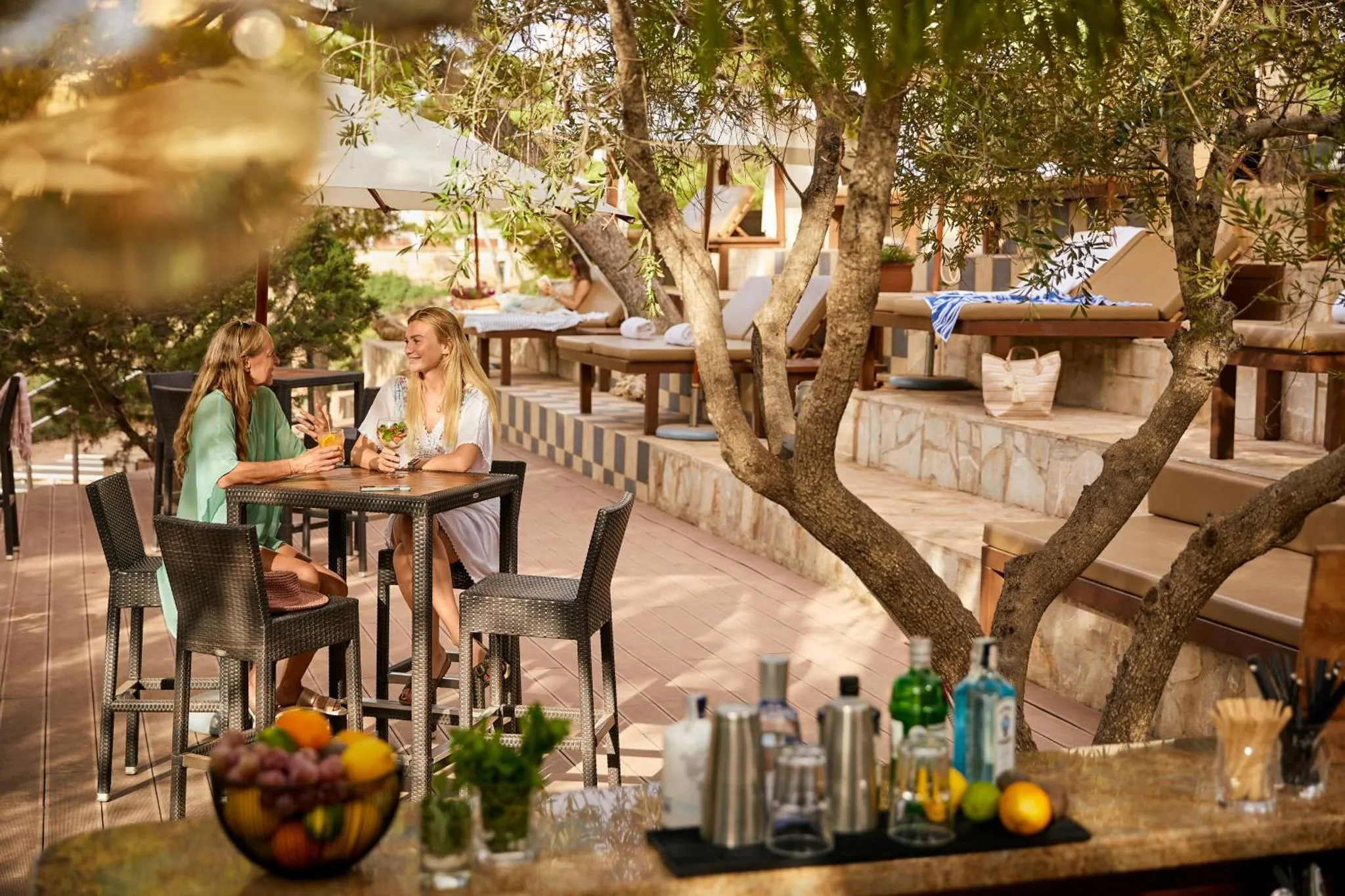 Deux femmes assises à une table de bar en terrasse, avec des boissons et des fruits.