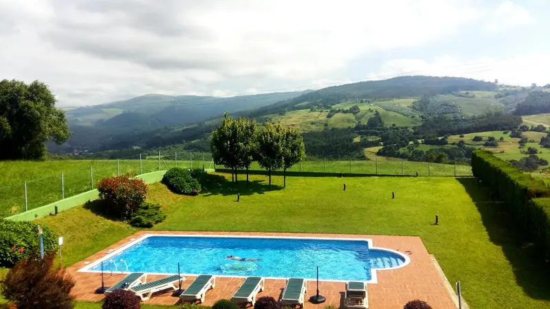 Une piscine rectangulaire avec des transats se trouve devant une pelouse verdoyante et des collines.
