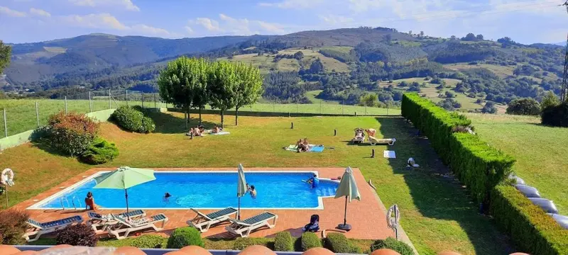 Une piscine rectangulaire avec des transats et des parasols, entourée d'une pelouse verdoyante et d'une haie soignée, avec des collines en arrière-plan.