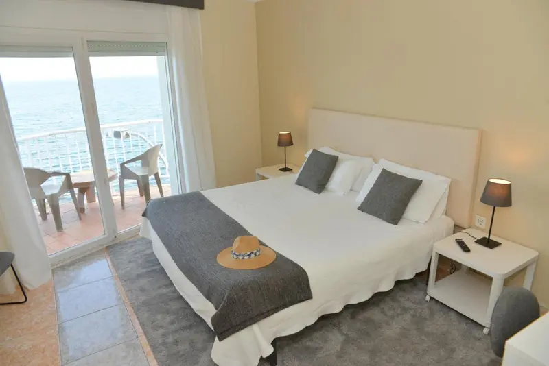 Chambre lumineuse avec un grand lit et une vue sur la mer depuis un balcon.