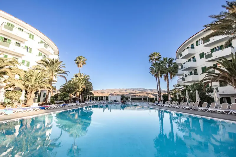 Une piscine turquoise reflète les bâtiments blancs et les palmiers sous un ciel bleu.
